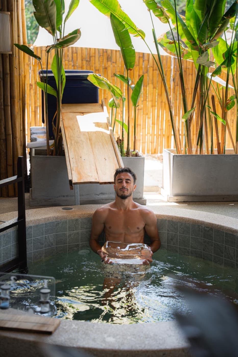 A person standing waist-deep in a wooden cold plunge tub of icy water, breathing steadily, at an outdoor sauna and ice bath retreat.